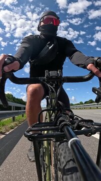 High speed vertical POV footage from a low angle of an athletic male cyclist in a black mask and a backward baseball cap sprinting fast on a gravel bike along a sunny highway.