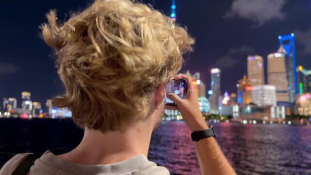 Summer Night at The Bund &ndash; Young Blond Man Standing on Waterfront with Smartphone, Reading, Scrolling and Taking Photos Against Skyscraper Skyline in Shanghai