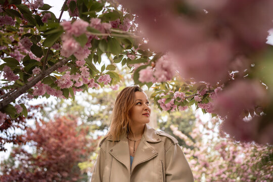 Middle aged woman in beige jacket stands amidst lush blooming pink cherry blossom branches during sunny spring day