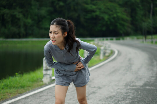 Medium full-length shot of an Asian female runner suffering from a painful side stitch cramp while training outdoors on a scenic road.