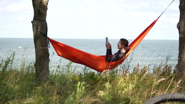 Cinematic sideways tracking shot moving from right to left past a young female traveler resting in a bright orange hammock and using her smartphone to take a photo of the sea.