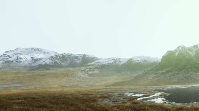 In the early morning, mountains with snowcaps rise above a grassy area. The sky is light and the landscape appears calm, showcasing natures simple beauty at sunrise.