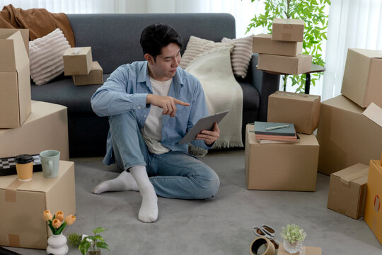 Young Asian man organizing home delivery business using digital tablet surrounded by shipping boxes