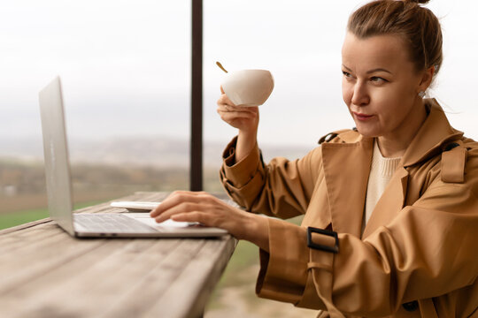Professional woman typing on laptop while holding coffee mug on scenic outdoor terrace overlooking landscape