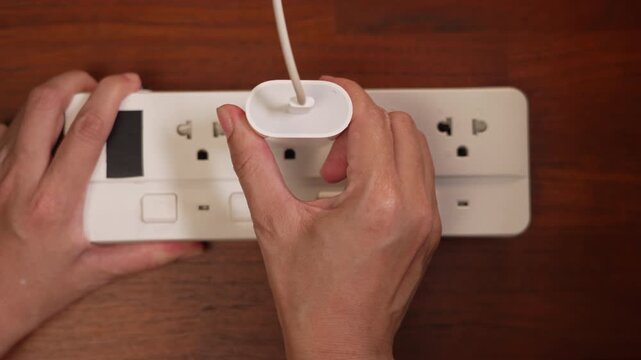 Top view of woman hands struggling to pull stuck adapter plug out of power strip on wooden desk, resistance concept.⁠
