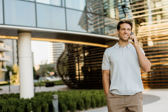 Smiling man walks along modern building while talking on phone