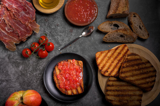 Toasted bread slices with cured Spanish ham, fresh tomatoes, and grated tomato sauce on dark surface, traditional healthy Mediterranean breakfast