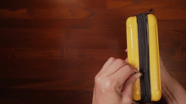 Centered top view of woman hands holding yellow hard case on wooden desk, failing to slide stuck zipper past halfway, deadlock concept.