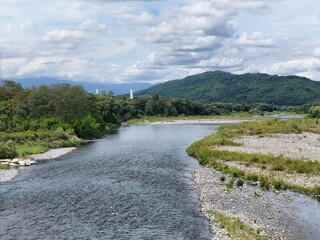 Serene river winds through lush green hills. Clouds drift above distant mountains, casting soft light. Shallow waters reveal smooth stones and vibrant grasses. Riverside banks host wildflowers © Masakazu Tokashiki