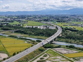 Aerial view of a curved highway bridging a river. Green fields stretch beyond the road's path. Distant mountains frame the horizon's edge. Clouds drift above the landscape's calm expanse © Masakazu Tokashiki