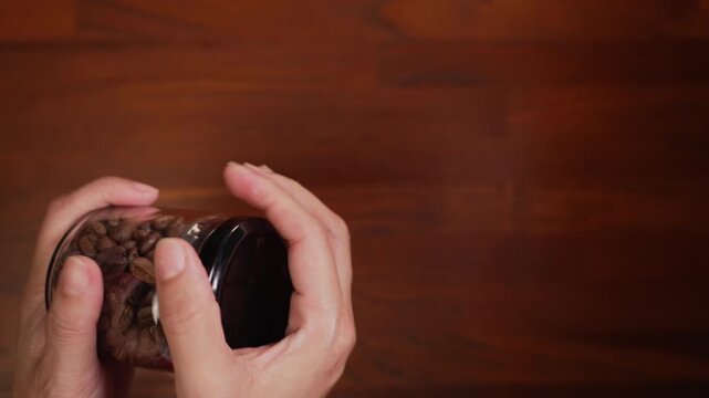 Top view on left side of woman hands reaching to pick up coffee bean glass jar on wooden desk, straining wrist to twist tight cap, failing, setting it back down, failure concept.⁠