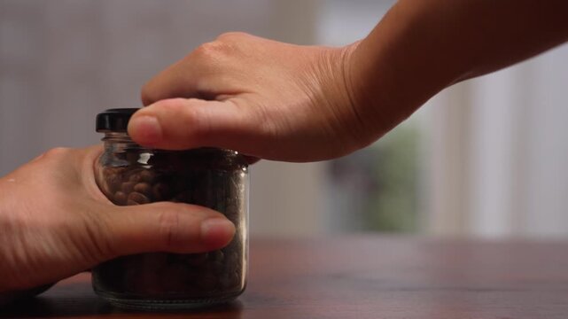 Close up on left side with copy space of woman hand supporting coffee glass jar on wooden desk by window, straining to twist tight cap until grip slips, obstacle concept.⁠
