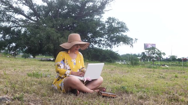Woman in Yellow Dress Working on Laptop While Sitting on Grass in Public Park