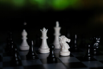 Chess piece strategy with white knight facing black opponent on checkered board creating dramatic tension in dimly lit setting © CoreRock