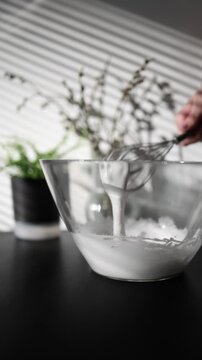 Vertical screen close up of a hand whisking a white egg mixture in a transparent glass bowl while preparing homemade desserts in a cozy kitchen