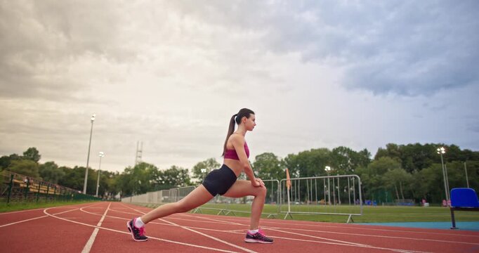 Young athletic woman in sportswear warming up and doing exercises on the stadium running track in the evening at sunset