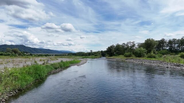 River flows gently through green landscape. Rocks line banks, grass grows wild and tall. Clouds drift lazily over distant mountains. Sky is bright, blue, with soft white patches