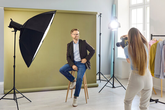 Photographer shoots model in studio portrait. Softbox lighting and seamless backdrop as a woman photographs a seated man for fashion branding. Clear concept of studio photoshoot with camera.