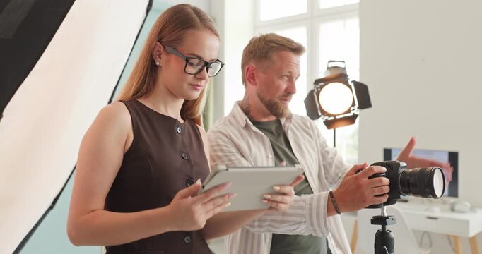 Photographer assistant camera studio lighting photoshoot teamwork production. They plan frames and review on a tablet, directing a shoot with tripod and lights. Clear teamwork and smart planning.