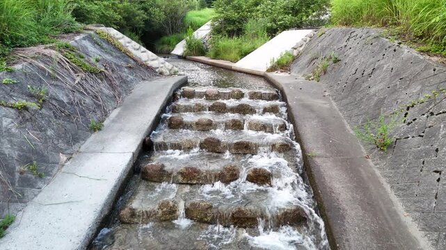 Water flows over stone steps in a man-made channel. Concrete edges line the stream's path. Green grass and trees frame the natural setting. Nature meets human engineering here