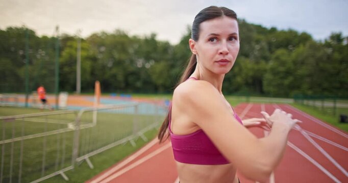 Young athletic woman in sportswear warming up and doing exercises on the stadium running track in the evening at sunset