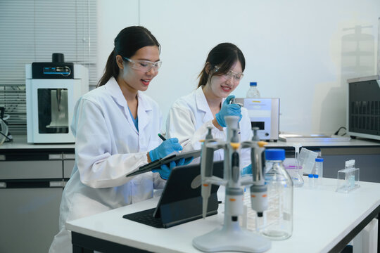 Two Asian Female Scientists Working Together in Modern Laboratory with Lab Equipment and Tablet
