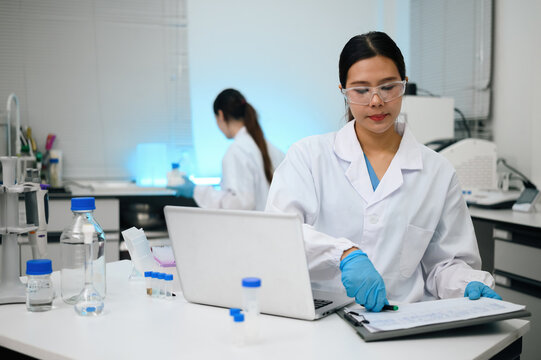Asian Female Scientist Working on Laptop in Modern Laboratory with Colleague in Background