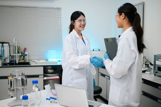 Two Asian Female Scientists Shaking Hands in Modern Laboratory, Wearing Lab Coats and Safety Goggles