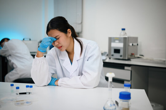 Stressed Asian Female Scientist Feeling Exhausted in Laboratory, Holding Head with Gloved Hand