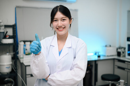 Smiling Young Asian Female Scientist in White Lab Coat Giving Thumbs Up in Modern Laboratory