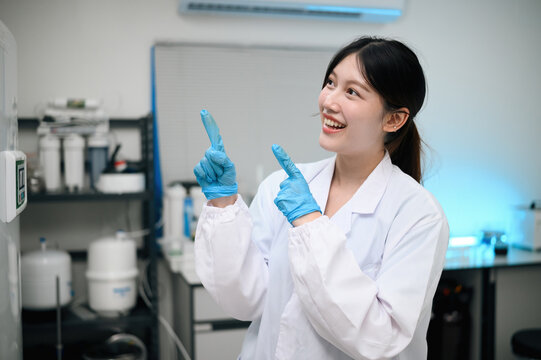 Happy Asian Female Scientist in White Lab Coat and Blue Gloves Pointing Up in Laboratory