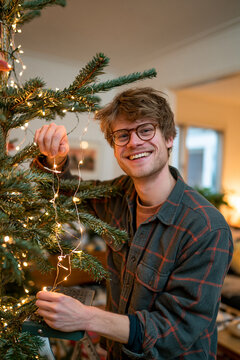Happy young man in glasses decorating Christmas tree with glowing garland lights at home during holiday season
