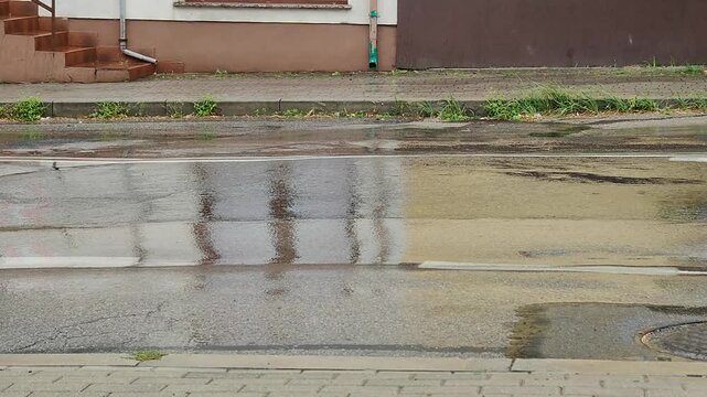 Deszcz padający na jezdnię, z kroplami wody tworzącymi odbicia budynk&oacute;w na mokrej nawierzchni.