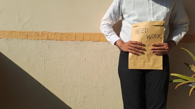 A job applicant holding a brown envelope with a handwritten need work text against a wall. Unemployment, career, and job hunting stock video footage.