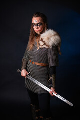 Fototapeta premium Young Viking woman with combat makeup on her face in chainmail with fur and a sword in her hands posing while standing against a dark background with blue backlighting.