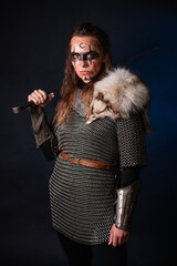 Fototapeta premium Young Viking woman with combat makeup on her face in chainmail with fur and a sword in her hands posing while standing against a dark background with blue backlighting.