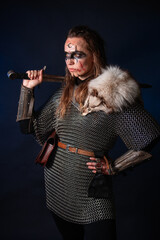 Fototapeta premium Young Viking woman with combat makeup on her face in chainmail with fur and a sword in her hands posing while standing against a dark background with blue backlighting.