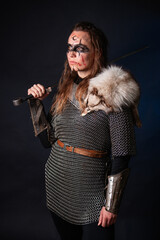 Fototapeta premium Young Viking woman with combat makeup on her face in chainmail with fur and a sword in her hands posing while standing against a dark background with blue backlighting.