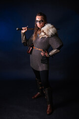 Fototapeta premium Young Viking woman with combat makeup on her face in chainmail with fur and a sword in her hands posing while standing against a dark background with blue backlighting.
