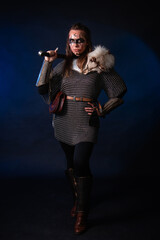 Fototapeta premium Young Viking woman with combat makeup on her face in chainmail with fur and a sword in her hands posing while standing against a dark background with blue backlighting.