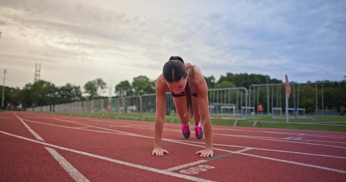Young athletic woman in sportswear warming up and doing exercises on the stadium running track in the evening at sunset