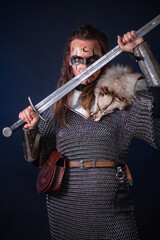 Fototapeta premium Young Viking woman with combat makeup on her face in chainmail with fur and a sword in her hands posing while standing against a dark background with blue backlighting.