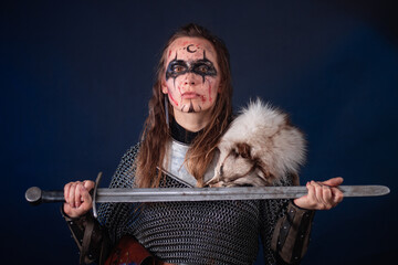 Fototapeta premium Young Viking woman with combat makeup on her face in chainmail with fur and a sword in her hands posing while standing against a dark background with blue backlighting.