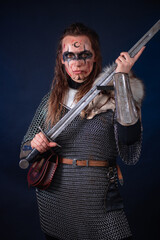 Fototapeta premium Young Viking woman with combat makeup on her face in chainmail with fur and a sword in her hands posing while standing against a dark background with blue backlighting.
