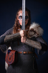 Fototapeta premium Young Viking woman with combat makeup on her face in chainmail with fur and a sword in her hands posing while standing against a dark background with blue backlighting.