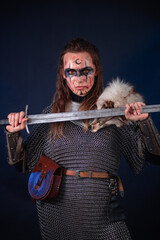 Fototapeta premium Young Viking woman with combat makeup on her face in chainmail with fur and a sword in her hands posing while standing against a dark background with blue backlighting.