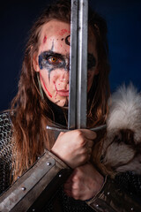 Fototapeta premium Young Viking woman with combat makeup on her face in chainmail with fur and a sword in her hands posing while standing against a dark background with blue backlighting.
