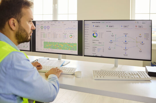 Engineer operator monitoring data on computer dashboards. In an industrial control room, dual screens show analytics as a safety worker manages operations. Strong concept of smart industry.