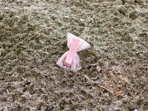 Close-up photograph of a discarded pink plastic wrapper and litter on sandy ground, highlighting environmental pollution.
