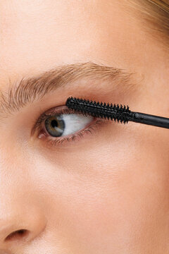 Attractive young woman applying mascara against a grey background.